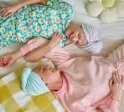 Two girls laying down on a bed, each wearing a Turbie twist hair towel. One is purple and one is aqua in color